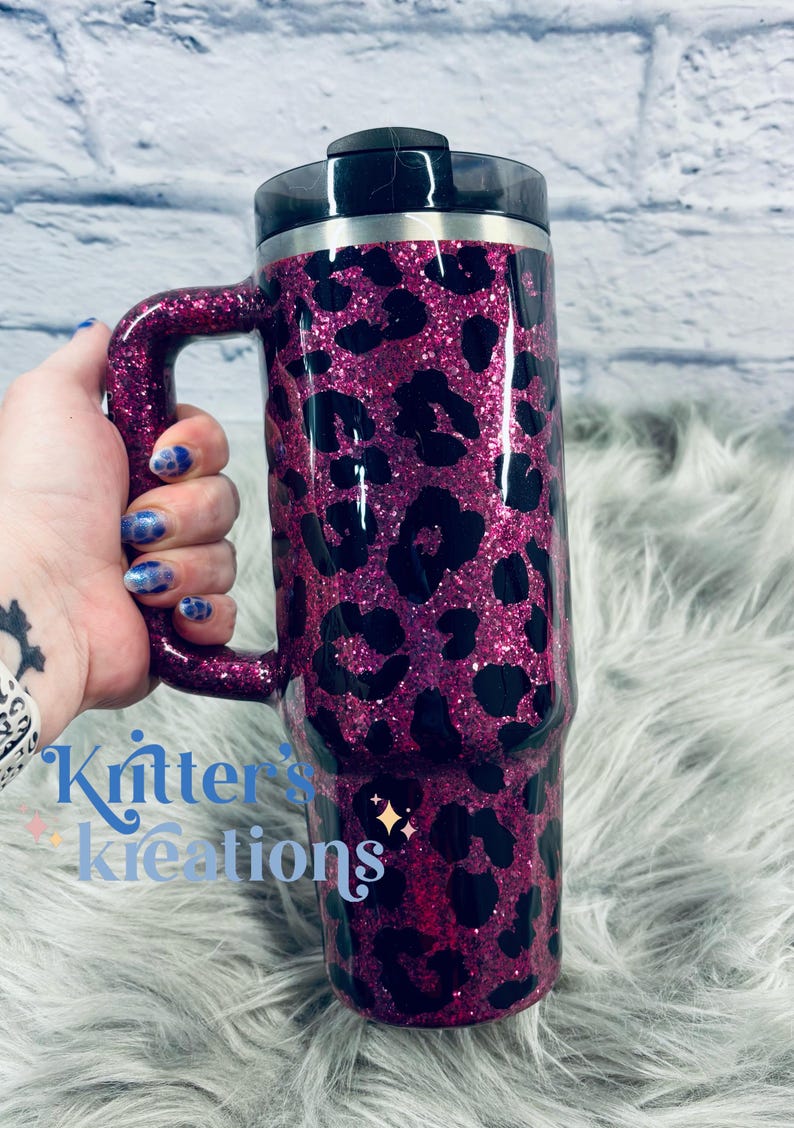 K&ouml;nnte beinhalten: Ein pinkfarbener Reisebecher mit Glitzer, schwarzem Deckel und Griff. Der Becher ist mit einem schwarzen Leopardenmuster versehen. Der Text "Kritter's Kreations" ist auf der Seite des Bechers zu sehen.