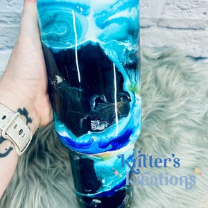 May include: A black and blue tumbler with a white and blue marbled design. The tumbler has a blue lid and a white rim. The tumbler is sitting on a blue and white fuzzy surface. The text "Knitter's Kreations" is visible on the bottom right of the image.