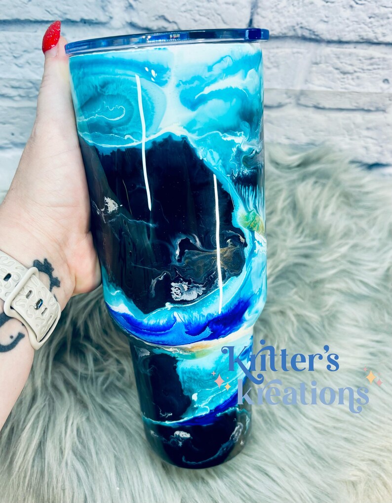 May include: A black and blue tumbler with a white and blue marbled design. The tumbler has a blue lid and a white rim. The tumbler is sitting on a blue and white fuzzy surface. The text "Knitter's Kreations" is visible on the bottom right of the image.