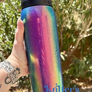 Peut inclure: Une bouteille d'eau cylindrique Owala avec une finition paillet&eacute;e multicolore vibrante passant par des tons de violet, rose et bleu sarcelle. La bouteille poss&egrave;de un bouchon noir et le texte "Kritter's Kreations" imprim&eacute; en bleu en bas.