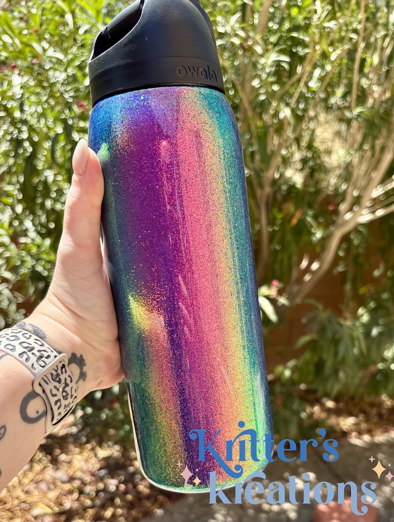 Peut inclure: Une bouteille d'eau cylindrique Owala avec une finition paillet&eacute;e multicolore vibrante passant par des tons de violet, rose et bleu sarcelle. La bouteille poss&egrave;de un bouchon noir et le texte "Kritter's Kreations" imprim&eacute; en bleu en bas.