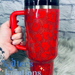 May include: A red glitter tumbler with a black lid and a red handle. The tumbler features a leopard print design in a darker shade of red. The tumbler is held by a hand.