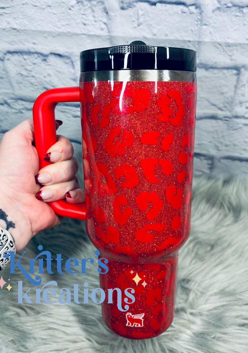 May include: A red glitter tumbler with a black lid and a red handle. The tumbler features a leopard print design in a darker shade of red. The tumbler is held by a hand.