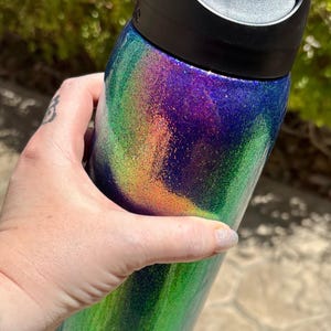 Peut inclure: Une bouteille d'eau cylindrique dot&eacute;e d'un couvercle en plastique noir et d'une finition scintillante aux d&eacute;grad&eacute;s tourbillonnants de violet, de vert et d'orange. La marque Owala est imprim&eacute;e en petites lettres noires pr&egrave;s de la base.