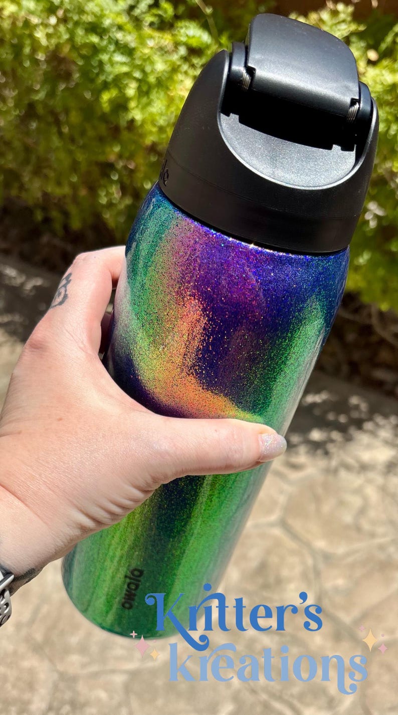 Peut inclure: Une bouteille d'eau cylindrique dot&eacute;e d'un couvercle en plastique noir et d'une finition scintillante aux d&eacute;grad&eacute;s tourbillonnants de violet, de vert et d'orange. La marque Owala est imprim&eacute;e en petites lettres noires pr&egrave;s de la base.