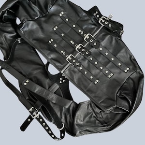 May include: A black leather garment featuring multiple adjustable straps with silver-toned metal buckles and rivets. The piece includes a central zipper closure and several horizontal strap overlays across the front, creating a structured design.