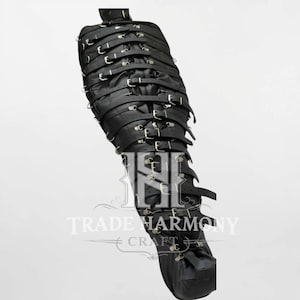 Könnte beinhalten: Schwarzer Leder-Fesselbeutel mit mehreren Riemen und Schnallen. Der Beutel ist so konzipiert, dass er den Körper umschließt, mit einem Halsverschluss und einem Kordelzug am Boden. Der Text "TRADE HARMONY CRAFT" ist sichtbar.
