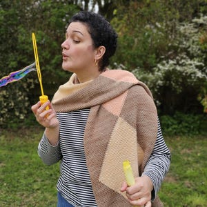 May include: A knitted scarf featuring a geometric pattern of large diamonds in shades of beige, tan, and peach. The scarf is draped over a black and white horizontal striped long-sleeve shirt and blue denim jeans, with yellow bubble wands.