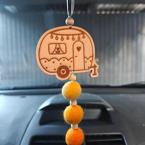 Könnte beinhalten: Holzanhänger in Wohnwagenform für das Auto, mit Herz- und Fensterdesign, an einer silbernen Schnur aufgehängt. Darunter sind drei Filzkugeln in Gelb- und Orangetönen mit weißen Perlen aufgefädelt. Der Anhänger hängt vor einem Armaturenbrett im Auto.