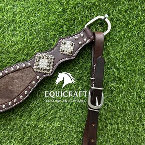 Puede incluir: Primer plano de un arn&eacute;s de caballo de cuero marr&oacute;n con herrajes plateados y adornos cuadrados decorativos. El arn&eacute;s presenta una secci&oacute;n de piel marr&oacute;n y blanca. El texto "EQUICRAFT LEATHER AND APPAREL" es visible.