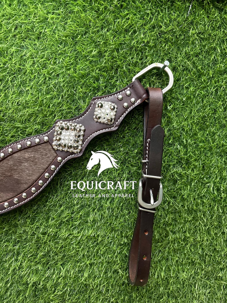 Puede incluir: Primer plano de un arn&eacute;s de caballo de cuero marr&oacute;n con herrajes plateados y adornos cuadrados decorativos. El arn&eacute;s presenta una secci&oacute;n de piel marr&oacute;n y blanca. El texto "EQUICRAFT LEATHER AND APPAREL" es visible.