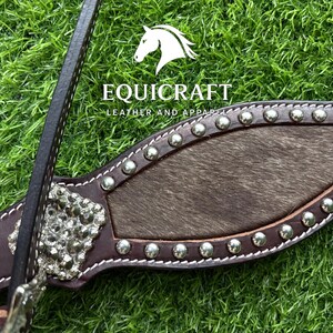 Puede incluir: Primer plano de un cabezal de caballo de cuero marr&oacute;n con tachuelas y adornos plateados. El cabezal presenta una secci&oacute;n central de piel oscura y el nombre de la marca "EQUICRAFT LEATHER AND APPAREL".