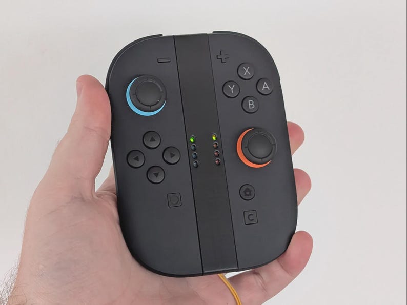 Puede incluir: Mando de juego port&aacute;til negro con joysticks azules y naranjas, botones direccionales y botones de acci&oacute;n. El mando tiene luces indicadoras verdes y se sostiene en una mano.