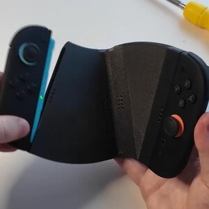 Puede incluir: Mando de videojuegos negro y azul con un joystick naranja y botones negros. El mando se sostiene con dos manos y parece estar en proceso de montaje. El mando tiene un dise&ntilde;o &uacute;nico.