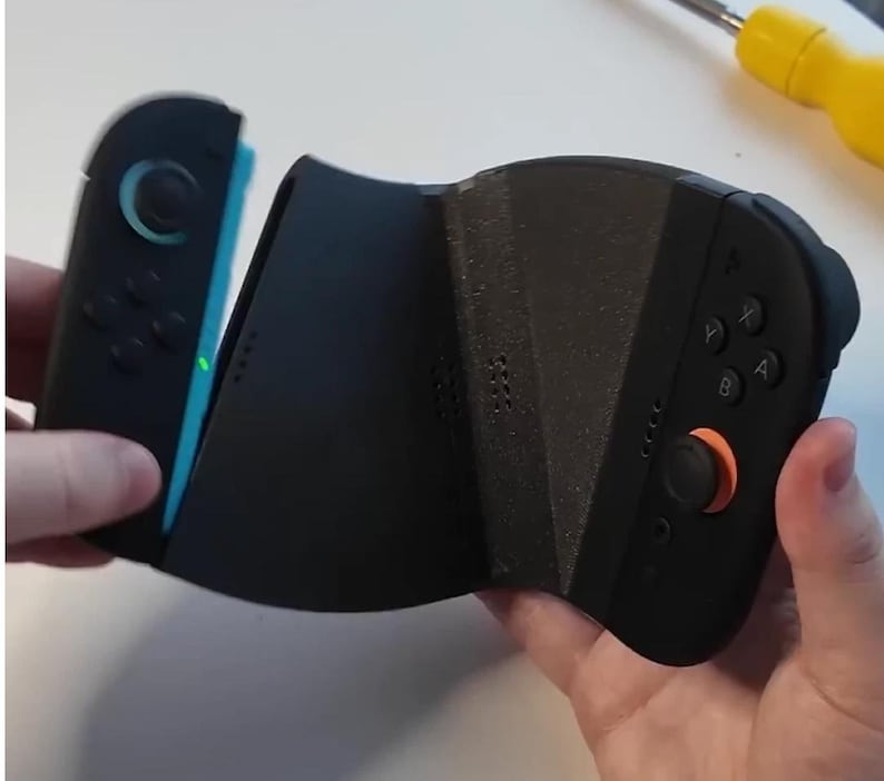 Puede incluir: Mando de videojuegos negro y azul con un joystick naranja y botones negros. El mando se sostiene con dos manos y parece estar en proceso de montaje. El mando tiene un dise&ntilde;o &uacute;nico.