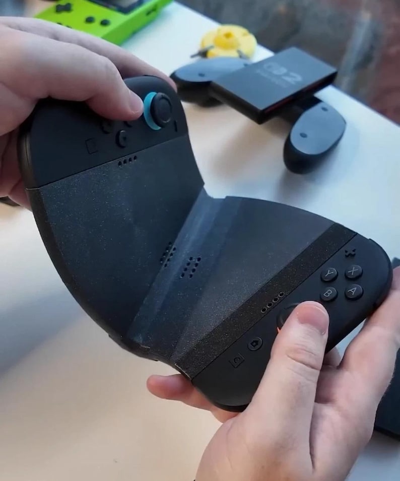 Puede incluir: Mando de juegos negro con un joystick azul y un agarre texturizado. El mando tiene botones etiquetados A, B, X e Y. El mando se sostiene con dos manos.