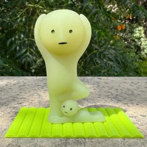 May include: A light green figurine of a person in a yoga pose, with arms raised and a small figure beneath. The figurine is on a bright green textured mat. The background is blurred green foliage.