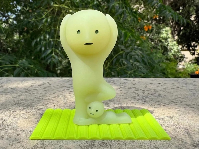 May include: A light green figurine of a person in a yoga pose, with arms raised and a small figure beneath. The figurine is on a bright green textured mat. The background is blurred green foliage.
