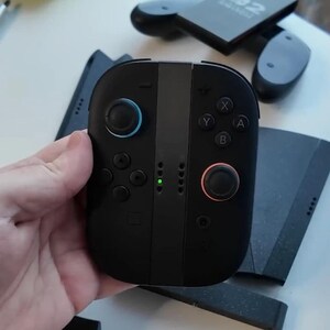 Puede incluir: Mando de juego negro con botones azules y rojos, y una franja central gris. El mando tiene una cruceta, botones etiquetados X, Y, A y B, y una peque&ntilde;a luz verde. Otros componentes del mando de juego son visibles en el fondo.