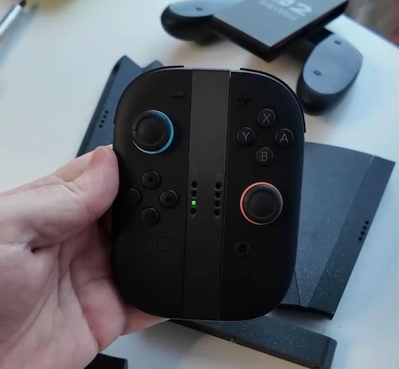 Puede incluir: Mando de juego negro con botones azules y rojos, y una franja central gris. El mando tiene una cruceta, botones etiquetados X, Y, A y B, y una peque&ntilde;a luz verde. Otros componentes del mando de juego son visibles en el fondo.