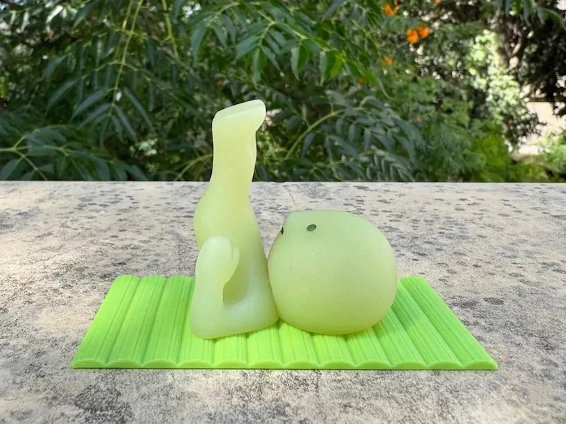May include: A light green, abstract sculpture with a rounded shape and a tall, curved form, resting on a textured, rectangular green mat. The background features lush green foliage.