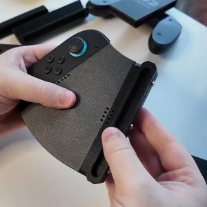 Puede incluir: Un accesorio de mando de juegos negro y gris con un agarre texturizado. El mando tiene varios botones y un joystick con un acento azul. Otros accesorios negros son visibles en el fondo.