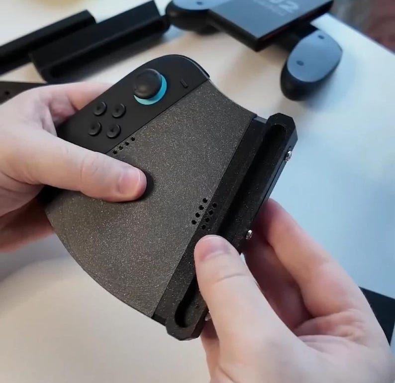 Puede incluir: Un accesorio de mando de juegos negro y gris con un agarre texturizado. El mando tiene varios botones y un joystick con un acento azul. Otros accesorios negros son visibles en el fondo.