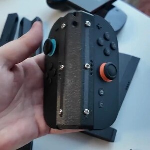Puede incluir: Mando de juegos port&aacute;til negro con joysticks, botones y tornillos azules y naranjas. El mando est&aacute; en la mano de una persona. Componentes negros adicionales son visibles en el fondo.