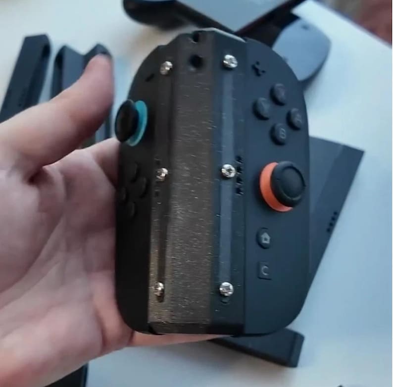 Puede incluir: Mando de juegos port&aacute;til negro con joysticks, botones y tornillos azules y naranjas. El mando est&aacute; en la mano de una persona. Componentes negros adicionales son visibles en el fondo.