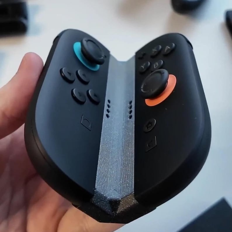 Puede incluir: Mando de juegos port&aacute;til negro con joysticks azules y naranjas y botones negros. El mando tiene un centro plateado y est&aacute; dise&ntilde;ado para sujetarse con dos manos. El mando tiene una cruceta, botones de acci&oacute;n y otros botones de control.