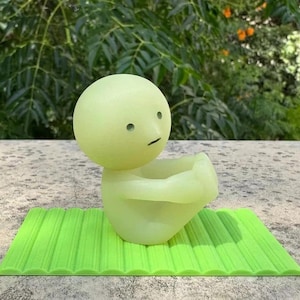 May include: A light green, cartoon-like figure with a round head and a small, curved mouth, sitting on a bright green, textured mat. The figure has two small, round eyes and is in a seated position. The background is blurred green foliage.