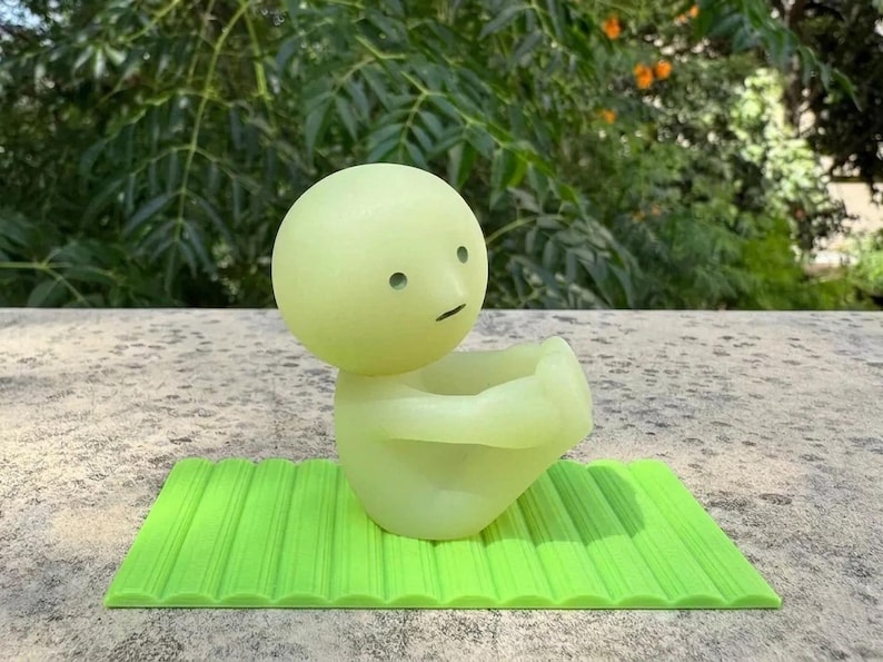 May include: A light green, cartoon-like figure with a round head and a small, curved mouth, sitting on a bright green, textured mat. The figure has two small, round eyes and is in a seated position. The background is blurred green foliage.