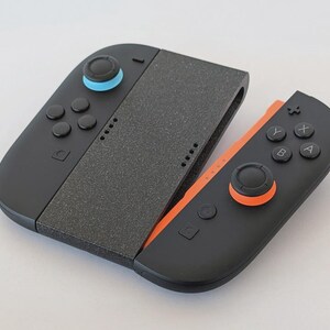 Puede incluir: Un mando de juego negro y naranja con una pieza central gris. El mando tiene botones, joysticks y un agarre texturizado. El joystick izquierdo es azul y el derecho es naranja.
