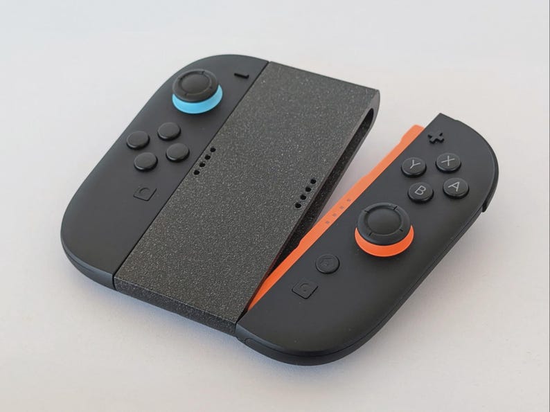 Puede incluir: Un mando de juego negro y naranja con una pieza central gris. El mando tiene botones, joysticks y un agarre texturizado. El joystick izquierdo es azul y el derecho es naranja.