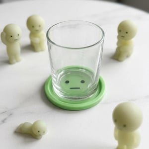 May include: A clear glass tumbler on a green coaster with a face design. Several small, pale yellow figurines are arranged around the glass. The scene has a minimalist aesthetic, with the figurines in different poses.
