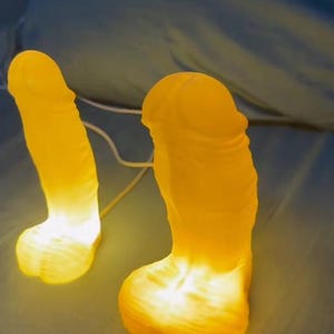 May include: Two illuminated yellow novelty lamps shaped like a phallus. The lamps are glowing with a warm light, creating a soft, inviting ambiance. The lamps are on a blue surface.