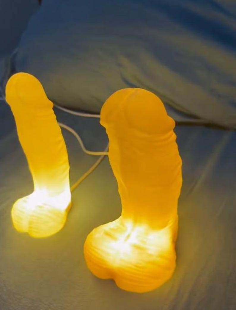 May include: Two illuminated yellow novelty lamps shaped like a phallus. The lamps are glowing with a warm light, creating a soft, inviting ambiance. The lamps are on a blue surface.