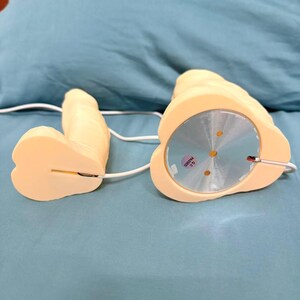 May include: A light beige sex toy with a rounded base and a phallic shape. The toy is connected by a white wire. The base has a silver disc with a QC sticker. The toy is on a blue surface.