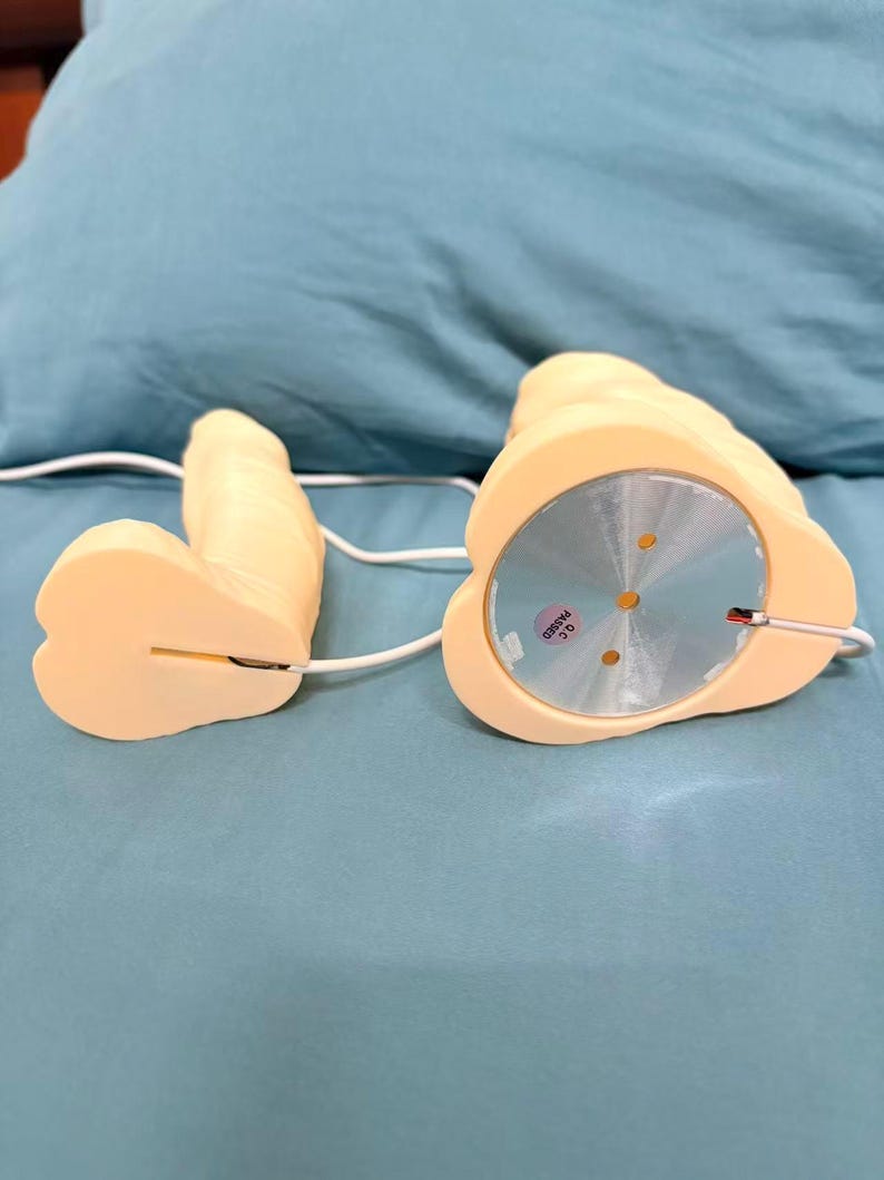 May include: A light beige sex toy with a rounded base and a phallic shape. The toy is connected by a white wire. The base has a silver disc with a QC sticker. The toy is on a blue surface.