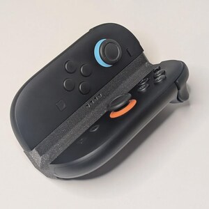 Puede incluir: Mando de juego negro con detalles azules y naranjas. El mando tiene varios botones y joysticks. El dise&ntilde;o es elegante y moderno, con un enfoque en la funcionalidad y la ergonom&iacute;a. El mando es probablemente para una consola de juegos.