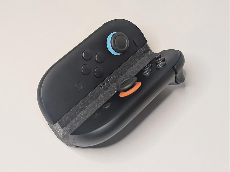 Puede incluir: Mando de juego negro con detalles azules y naranjas. El mando tiene varios botones y joysticks. El dise&ntilde;o es elegante y moderno, con un enfoque en la funcionalidad y la ergonom&iacute;a. El mando es probablemente para una consola de juegos.