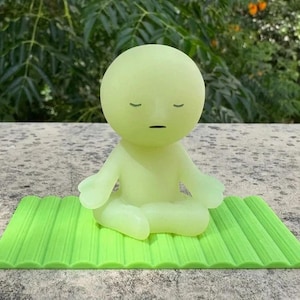 May include: A light green figurine in a seated yoga pose, with closed eyes and a neutral expression, sits on a bright green textured mat. The figurine is a solid color and appears to be made of a smooth material. The background is blurred.