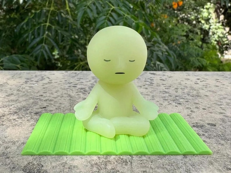 May include: A light green figurine in a seated yoga pose, with closed eyes and a neutral expression, sits on a bright green textured mat. The figurine is a solid color and appears to be made of a smooth material. The background is blurred.