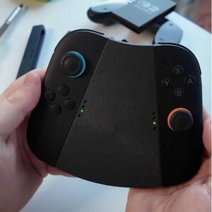 Puede incluir: Mando de juegos negro con joysticks azules y naranjas, y luces indicadoras verdes. El mando tiene botones etiquetados X, Y, A y B. El mando se sostiene con dos manos.