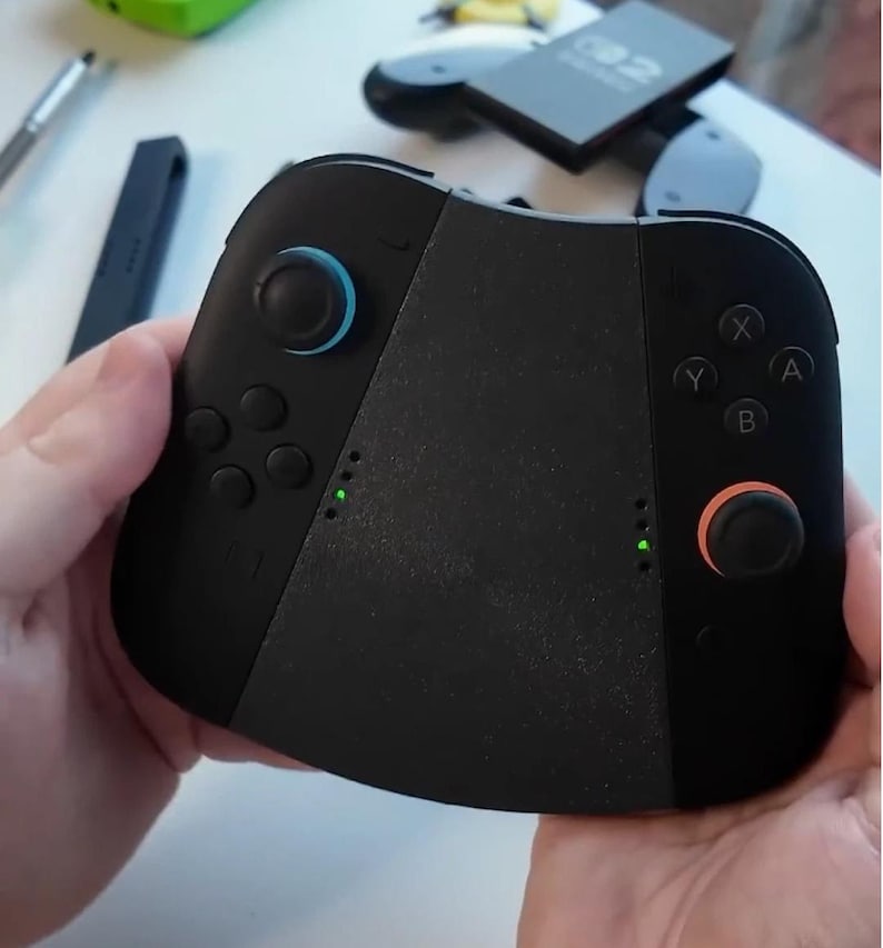 Puede incluir: Mando de juegos negro con joysticks azules y naranjas, y luces indicadoras verdes. El mando tiene botones etiquetados X, Y, A y B. El mando se sostiene con dos manos.
