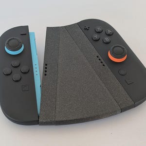Puede incluir: Mando de juego negro y azul con agarre gris. El mando tiene botones con detalles en naranja y azul. El mando est&aacute; dise&ntilde;ado para ser utilizado con una consola de juegos.