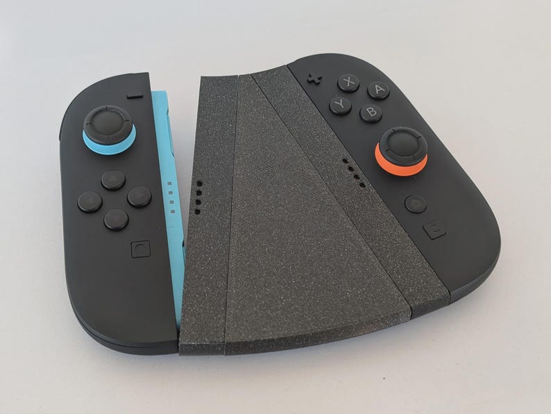 Puede incluir: Mando de juego negro y azul con agarre gris. El mando tiene botones con detalles en naranja y azul. El mando est&aacute; dise&ntilde;ado para ser utilizado con una consola de juegos.