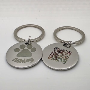 Könnte beinhalten: Zwei runde, silberne Hundeanhänger mit Schlüsselringen. Ein Anhänger zeigt einen Pfotenabdruck und den Namen "Ashley". Der andere Anhänger zeigt einen QR-Code. Beide Anhänger sind aus glänzendem Metall.