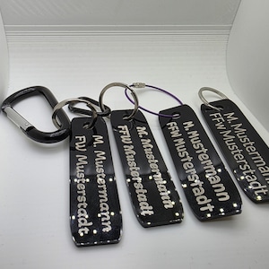 Könnte beinhalten: Eine Sammlung schwarzer Schlüsselanhänger mit silbernen Schlüsselringen und einem schwarzen Karabinerhaken. Jeder Schlüsselanhänger trägt den Text "M. Mustermann FFW Musterstadt" in silberner Schrift. Die Schlüsselanhänger sind rechteckig mit abgerundeten Kanten.