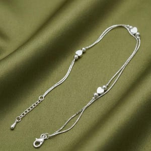 May include: A delicate silver anklet with a double strand design. The anklet features small heart-shaped charms and tiny round beads. The adjustable chain has a lobster clasp closure. The anklet is displayed on a green satin fabric.